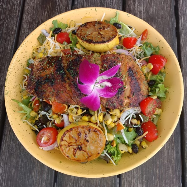 Grilled chicken salad with corn, tomatoes, black beans, cheese, and grilled lemon halves, garnished with an orchid.
